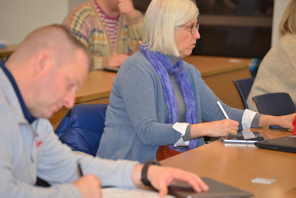 Adult students sit a tables in a classroom and write.