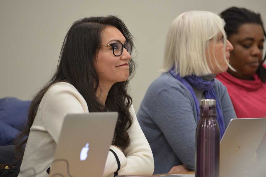 Three adult students sit at tables in a classroom with laptops open in front of them.